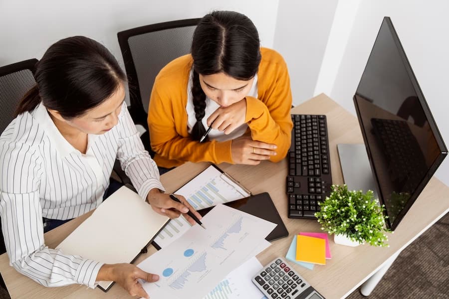 A person in a plaid shirt gestures towards floating bar and pie charts overlaid on printed financial reports. A laptop, tablet, and calculator are visible.