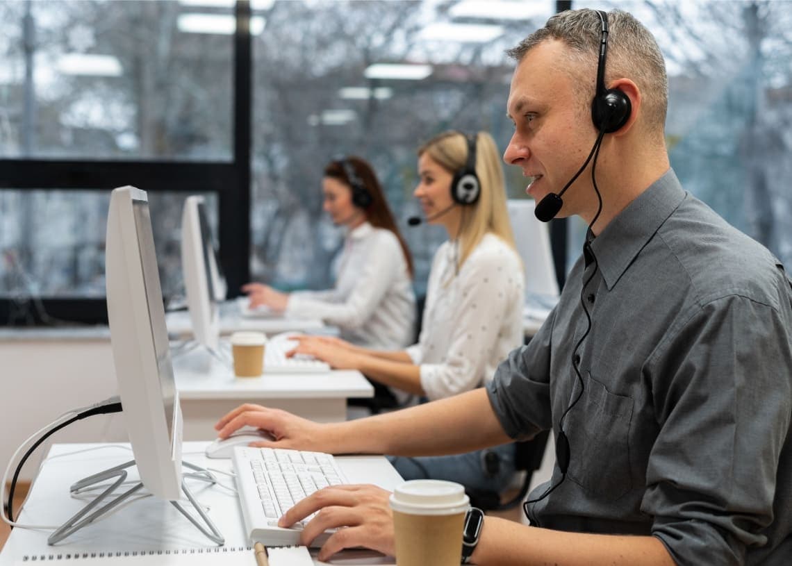 Three customer service representatives wearing headsets work at computers in a bright, modern office. They appear focused and professional.