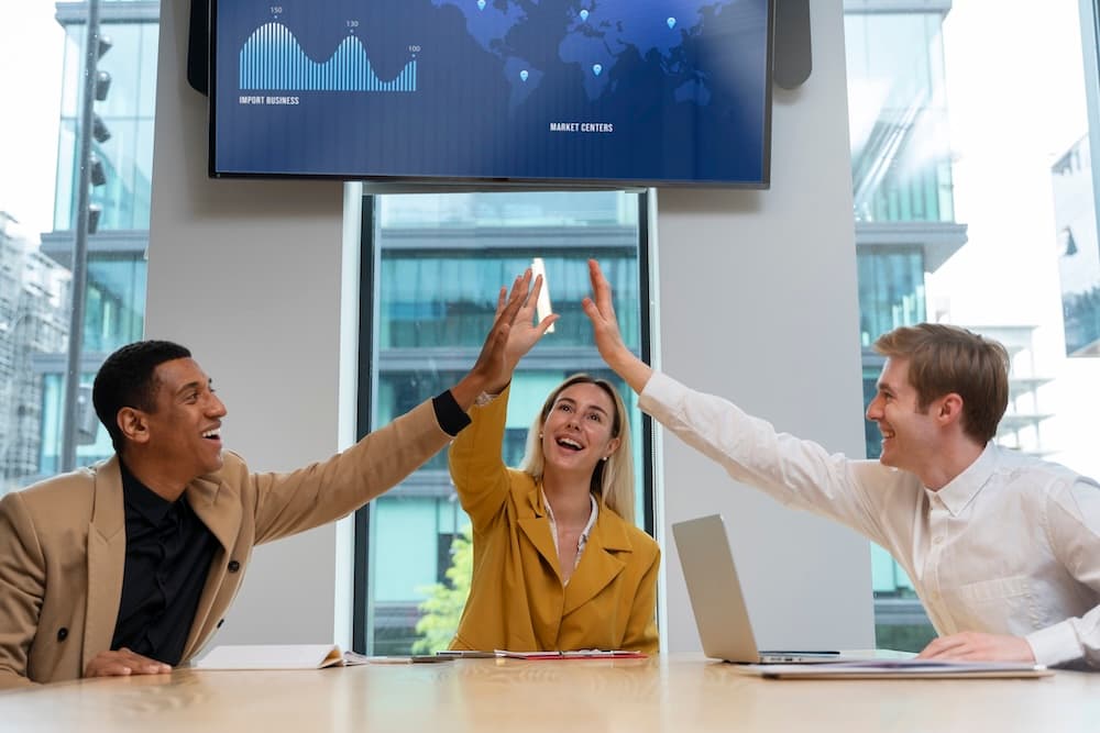 Three colleagues smiling and high-fiving around a conference table, with a laptop open. Three colleagues smiling and high-fiving around a conference table, with a laptop open.