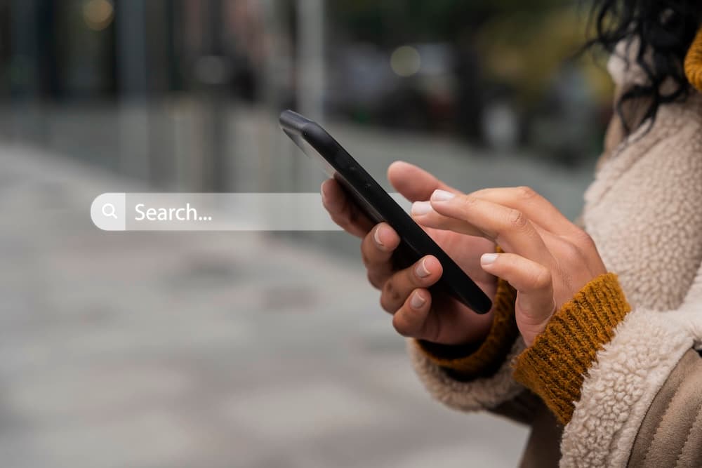 A person in a tan coat uses a smartphone outdoors. A translucent search bar with the word 'Search...' overlays the image. A person in a tan coat uses a smartphone outdoors. A translucent search bar with the word 'Search...' overlays the image.