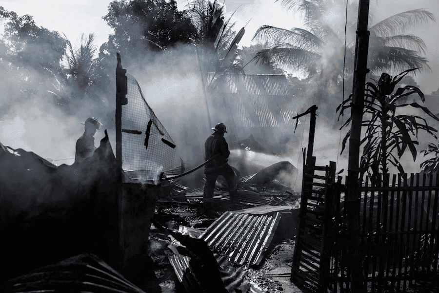 Firefighters battling a smoky blaze amid debris and ashes, silhouetted against hazy sunlight and palm trees. The scene conveys urgency and intensity. Firefighters battling a smoky blaze amid debris and ashes, silhouetted against hazy sunlight and palm trees. The scene conveys urgency and intensity.