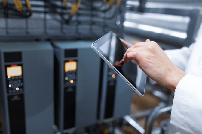A person in a white coat uses a tablet in an industrial setting with control panels and cables. The scene conveys a sense of technology and precision. A person in a white coat uses a tablet in an industrial setting with control panels and cables. The scene conveys a sense of technology and precision.